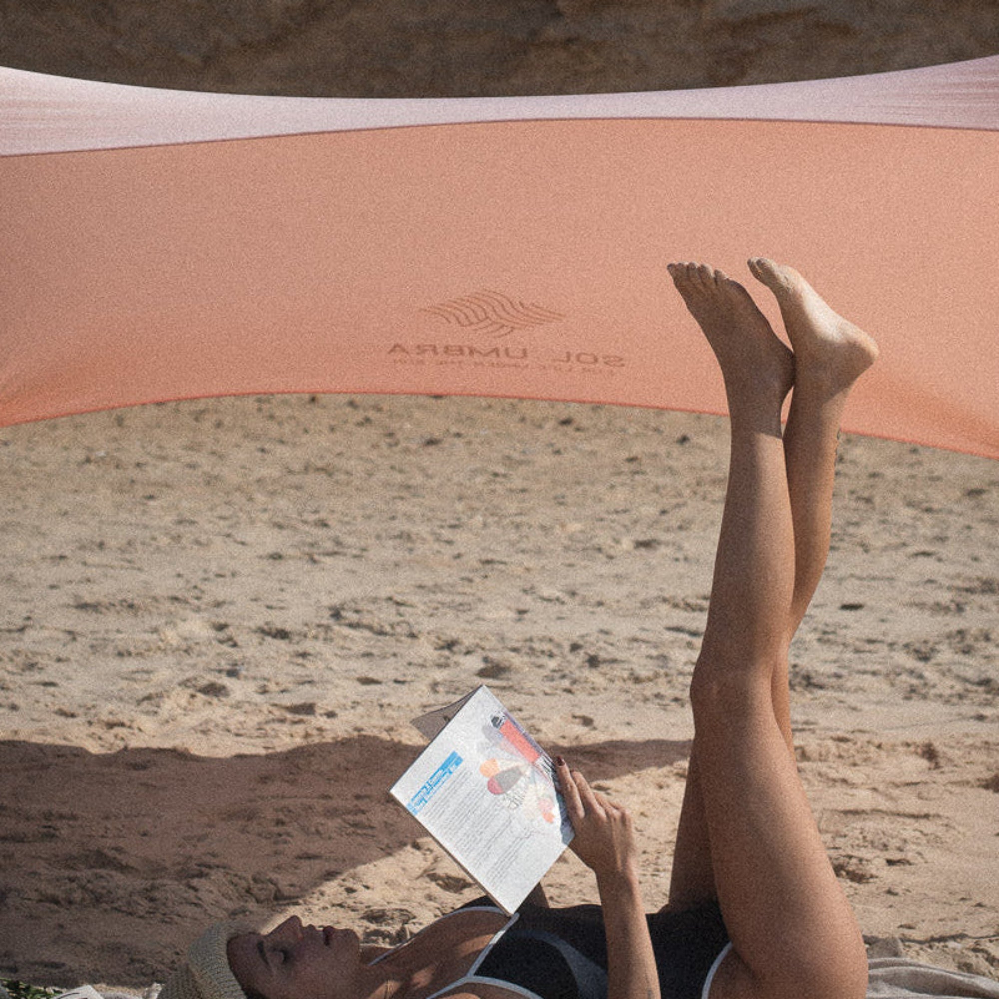 Person lying on a towel reading a book under a pink canopy on a sandy beach.